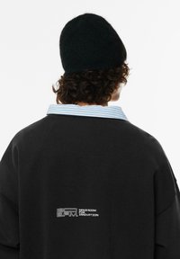 Černá mikina s límečkem v světle modrých pruzích, se bílým logem na zádech. Měkká textura a oversized střih. Černá pletená čepice.