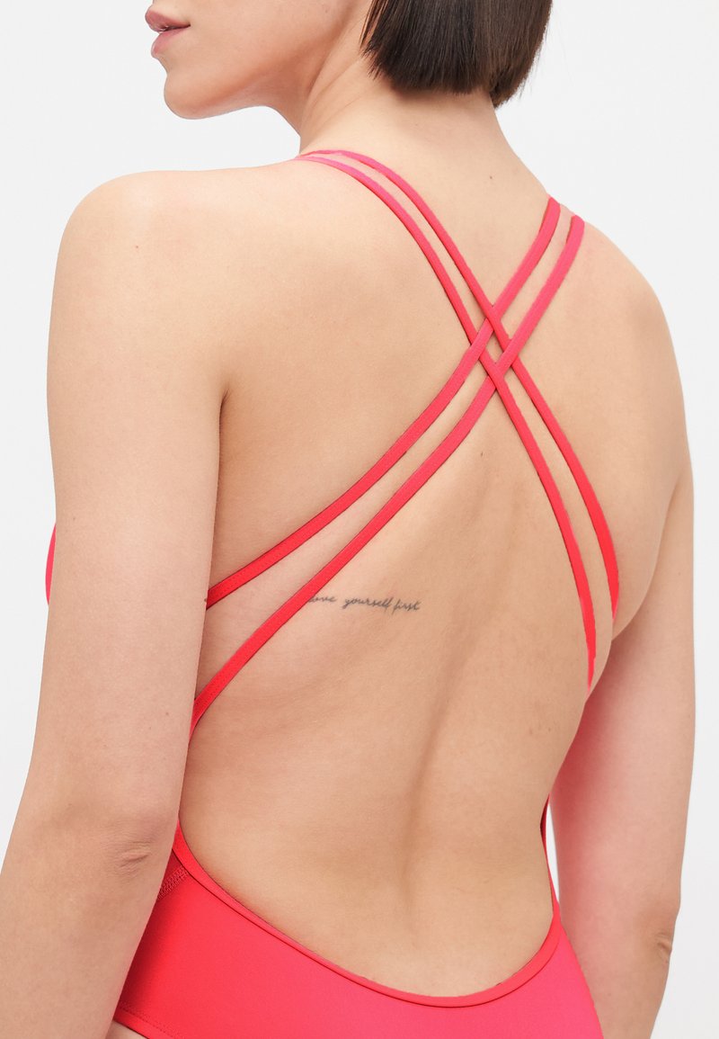 Bañador rojo con tirantes entrelazados en la espalda, textura suave y escote redondeado, con un pequeño tatuaje visible en la parte baja de la espalda.