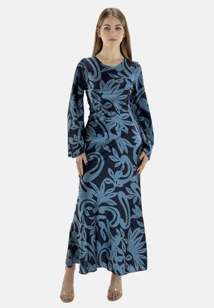 Frau in einem langen, figurbetonten dunkelblauen Kleid mit großen hellblauen Blumenmustern und langen Ärmeln, die vor einem einfarbigen Hintergrund steht.