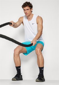 Bijela bezrukavna Nike majica, tirkizne kratke hlače, crni tenisice, držeći crne trening konopce, s jednostavnom bijelom pozadinom.