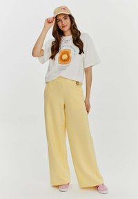 Bílý tričko s grafickým potiskem palačinky, žluté široké kalhoty a béžová kšiltovka. Růžové sneakers s bílou podrážkou. Pohodlný, volný střih.