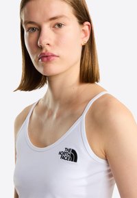 Fiatal nő egyenes barna hajjal, kis arany karika fülbevalókkal és egy fehér The North Face ujjatlan felsőben, egyszínű világos háttér előtt.