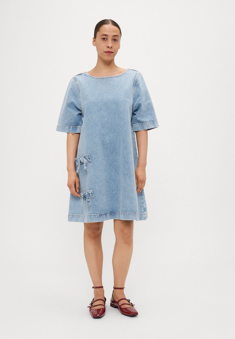 Vestido de ganga em azul claro, com mangas curtas, decote redondo, duas laços decorativos nos bolsos e barra desfeita. Usado com sapatos de tornozelo vermelhos.
