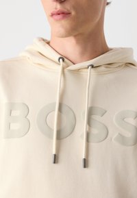 Sudadera color crema con el logo "BOSS" en relieve en un tono sutil, cordones con puntas metálicas y una textura suave y lisa.
