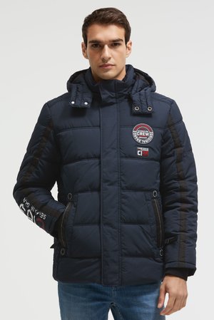 Uomo che indossa una giacca trapuntata blu navy con toppe sul petto e sulla manica, con le mani nelle tasche e jeans blu, su sfondo semplice.