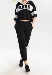 Persoon in een zwart-wit Puma cropped shirt, zwarte Puma joggingbroek en zwarte sportschoenen, staand met handen in de zakken.