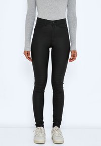 Zwarte high-waisted skinny jeans met een gladde textuur, vijf zakken en een ritsluiting. Gecombineerd met witte sneakers en een grijze lange mouwen top.