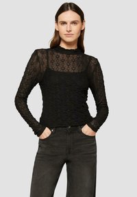 Zwarte kant lange mouwen top met een hoge hals en bloemenpatroon, gelaagd over een effen zwarte tanktop; gecombineerd met donkere jeans.