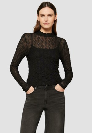 Haut à manches longues en dentelle noire avec un col haut et un motif floral, superposé sur un débardeur noir uni ; associé à un jean foncé.