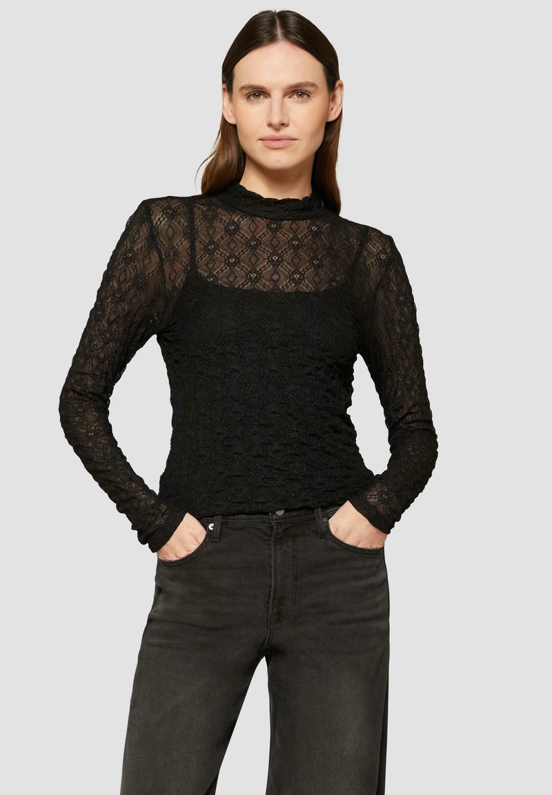 Zwarte kant lange mouwen top met een hoge hals en bloemenpatroon, gelaagd over een effen zwarte tanktop; gecombineerd met donkere jeans.