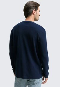 Mann trägt ein langärmliges marineblaues Hemd und hellblaue Jeans, schaut von hinten gegen einen einfarbigen hellgrauen Hintergrund.