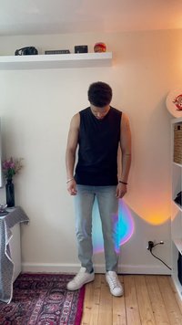 Top sem mangas preto, jeans azul claro e sapatilhas brancas; pose de pé com chão de madeira texturizada e iluminação ambiente suave.