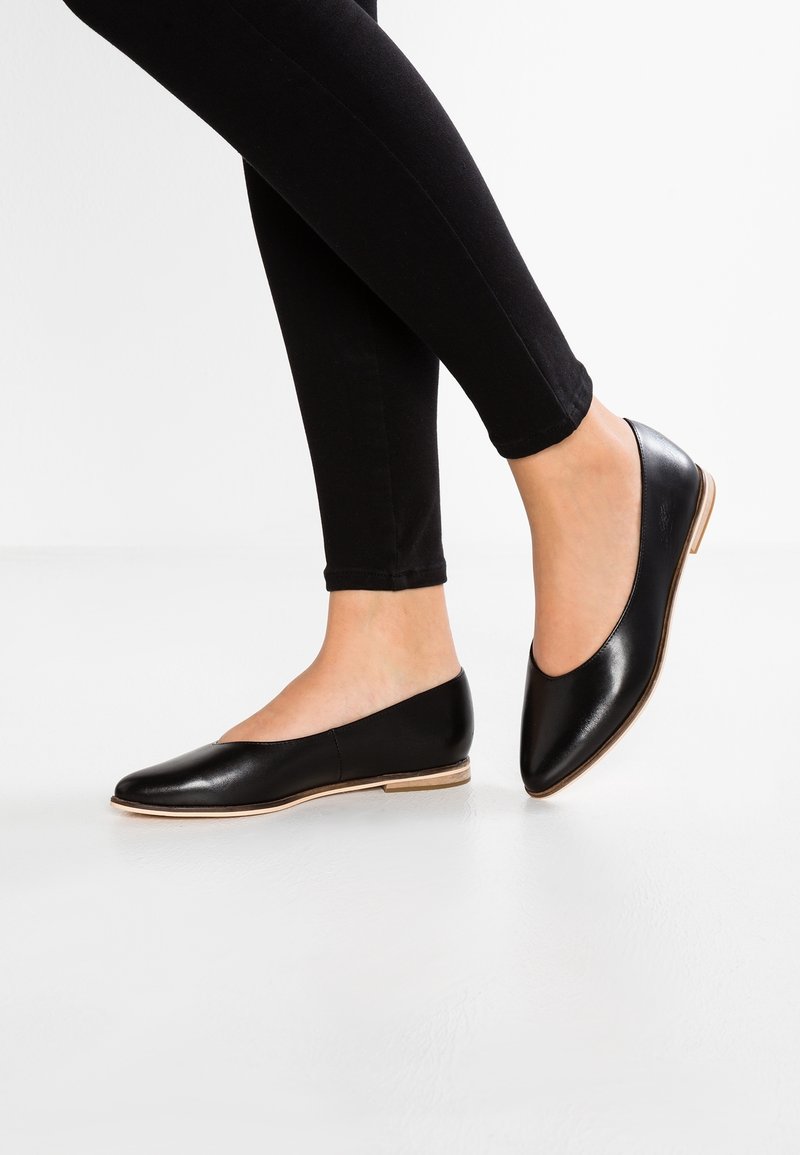 Ballerines noires en cuir, à bout pointu, avec une surface lisse et un design minimaliste. Talon fin de couleur claire, portées avec des leggings noirs.