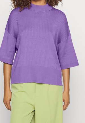 Femme portant un pull violet côtelé à manches courtes et un pantalon vert clair sur un fond uni.