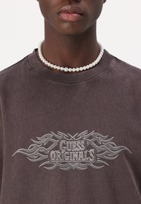 Pelēks kokvilnas T-krekls ar liesmu dizainu un "GUESS ORIGINALS" tekstu, ar apaļu kakla izgriezumu. Apvienots ar pērļu kaklarotu ap kaklu.