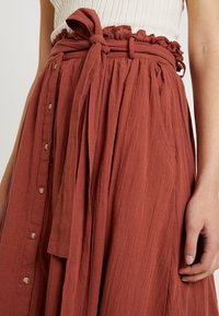 Woman wearing rust-colored gathered skirt with fabric belt tied in bow and button details along the front, white ribbed top partially visible.