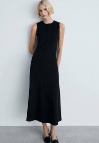 Schwarzes ärmelloses Midikleid mit geradem Schnitt, das mit einer Vordernaht und glatter Textur versehen ist, kombiniert mit schwarzen Slippers.