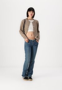 Cardigan castanho com botões, top branco cropped, jeans azuis e ténis pretos. O conjunto é casual e ajustado ao corpo.