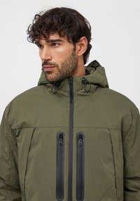 Giacca verde oliva con zip frontale, cappuccio e due tasche verticali con zip, caratterizzata da accenti neri e una texture liscia opaca.
