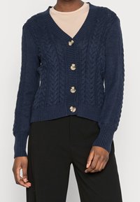 Mörkblå stickad kavaj med V-hals, med kabelmönster, ribbad nederkant och sex runda, glänsande knappar framtill.