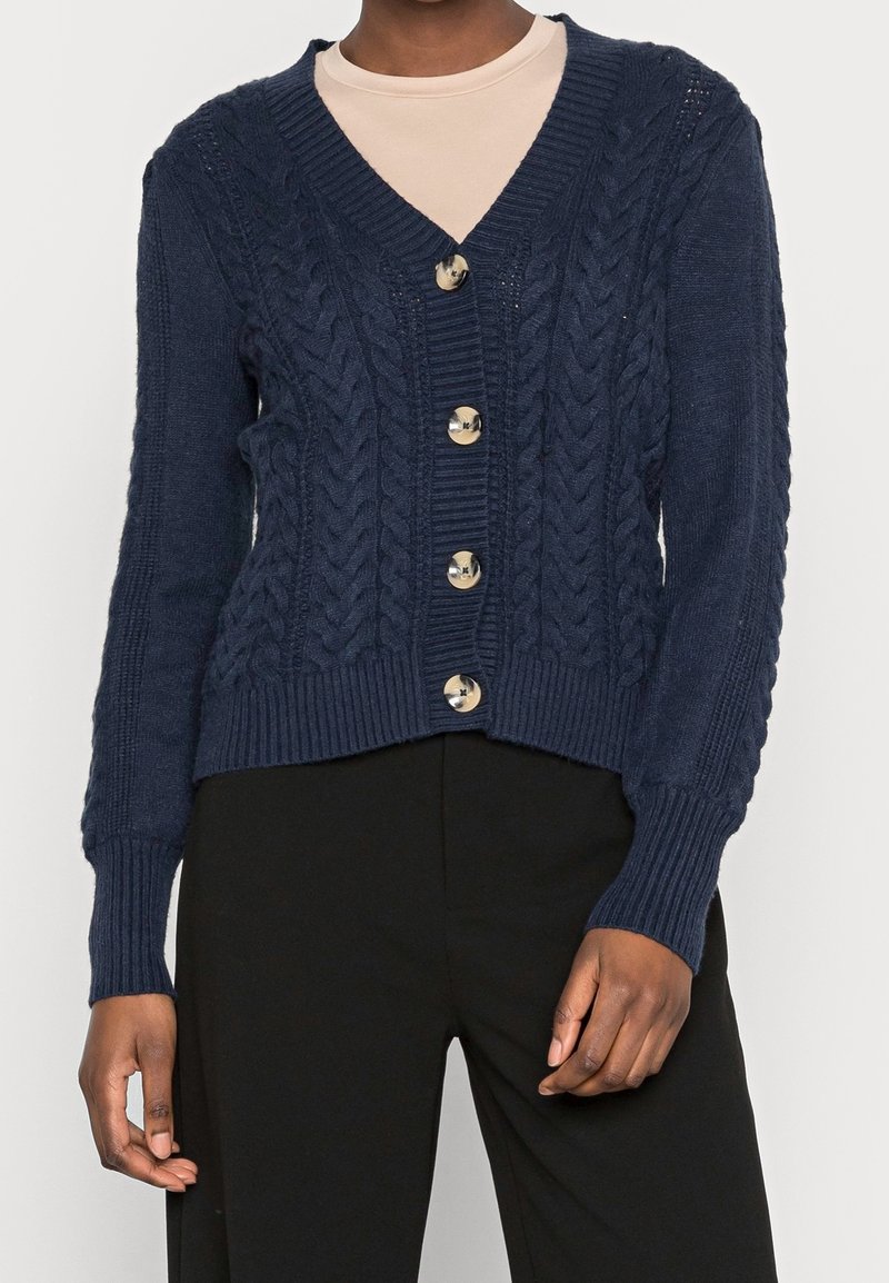 Mörkblå stickad kavaj med V-hals, med kabelmönster, ribbad nederkant och sex runda, glänsande knappar framtill.