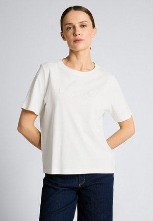 Młoda kobieta o jasnej karnacji i brązowych włosach związanych do tyłu, ubrana w prostą białą koszulkę z krótkim rękawem i ciemnoniebieskie dżinsy, stojąca na szarym tle.