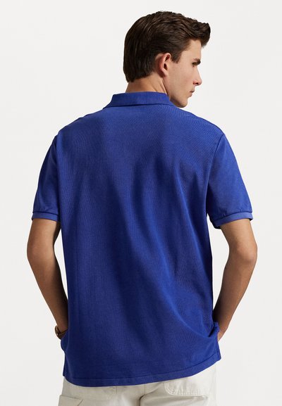 Tricou polo de culoare albastră solidă, cu mâneci scurte și guler clasic. Materialul pare texturat, cu o croială regulară și slituri laterale pentru confort.