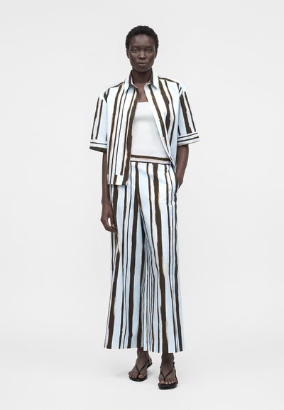 Γυναίκα με κοντομάνικο πουκάμισο με κάθετες ρίγες σε ανοιχτό μπλε και σκούρο καφέ χρώμα και ταιριαστό παντελόνι με φαρδιά πόδια, λευκό αμάνικο τοπ και μαύρα σανδάλια.