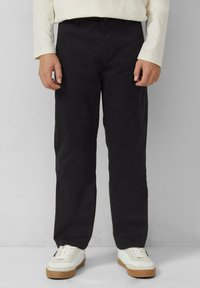 Person, der schwarze gerade geschnittene Hosen, ein beiges langärmeliges Hemd und weiße Sneakers mit braunen Sohlen trägt, steht vor einem einfarbigen Hintergrund.