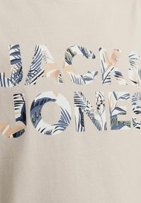 Beige puuvillane T-särk, millel on julge tekst "JACK & JONES", mis on täidetud troopilise lehe mustriga roheline, sinine ja persikatooni varjundites.
