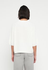 Άσπρο πλεκτό τοπ με κοντά raglan μανίκια, άνετη εφαρμογή και ριμπ λαιμόκοψη. Φορεμένο με ανοιχτό γκρι ψηλόμεσα παντελόνια.