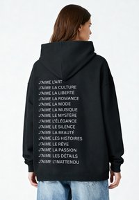 Schwarze Kapuzenjacke mit weißen Texten auf dem Rücken, die "J�’aime"-Aussagen zeigen. Weicher Stoff, oversized Schnitt und Kapuze mit Kordelzug. Lässiges Design.