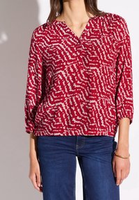 Kobieta ubrana w długą bluzkę z czerwono-białym wzorem, z marszczonymi rękawami, oraz niebieskie dżinsy, stojąca z ramionami swobodnie opuszczonymi wzdłuż ciała.