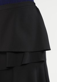 Lagererad svart kjol med slät tyg och asymmetrisk nederkant mot en blank ljus bakgrund.