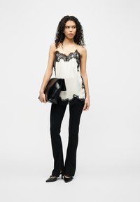 Γυναίκα με σγουρά μαλλιά, φορούσα λευκό καλοκαιρινό τοπ με δαντέλα, μαύρο παντελόνι καμπάνα, μυτερά παπούτσια, κρατώντας μεγάλη μαύρη δερμάτινη τσάντα.