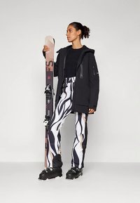 Frau in schwarzer Jacke, mit schwarz-weißen, gemusterten Skihosen und Skischuhen, die ein aufrechtes Paar Skier hält.