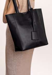 Sac fourre-tout en cuir noir avec deux longues poignées, forme structurée, détails de couture subtils et une étiquette rectangulaire.