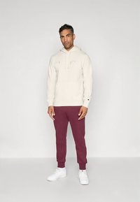 Sweatshirt creme com capuz, bolso frontal e cordão, combinada com calças de treino borgonha. Ambas feitas de tecido macio; sapatilhas brancas completam o conjunto.