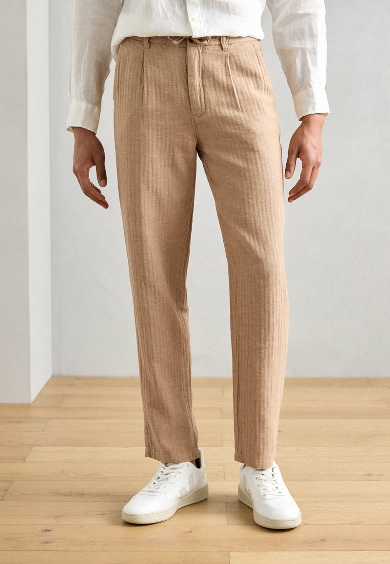 Lysbrune, teksturerede bukser med lodrette striber, der har en afslappet pasform og elastisk talje. Sæt med hvide sneakers.