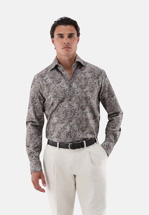 Homme portant une chemise à manches longues à motif floral rentrée dans un pantalon blanc avec une ceinture noire, debout devant un fond uni.
