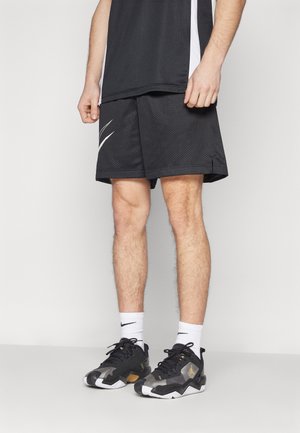 Männliche Beine mit schwarzen Sportshorts, weißen Nike-Socken und schwarz-weißen Nike-Sneakern, die vor einem schlichten hellen Hintergrund stehen.