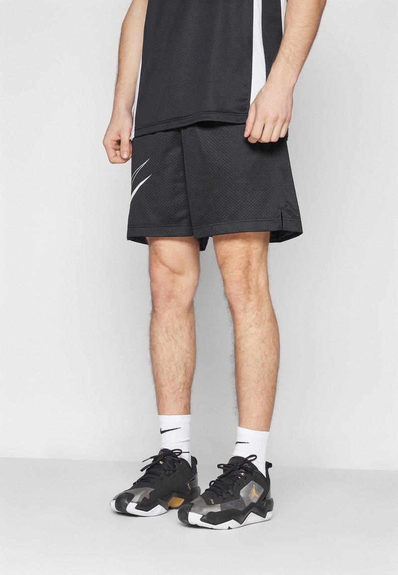 Jambes masculines portant un short de sport noir, des chaussettes blanches Nike et des baskets Nike noires et blanches, posées devant un fond clair uni.