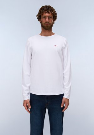 Mand med krøllet hår og skæg iført en enkel hvid langærmet skjorte med et lille rødt logo på brystet og mørkeblå jeans.