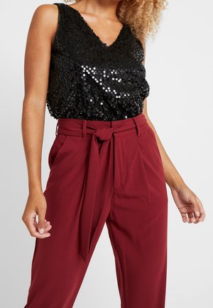 Kobieta w czarnej bezrękawnikowej bluzce z cekinami i wysokich bordowych spodniach z przewiązanym paskiem, stojąca na jednolitym tle.