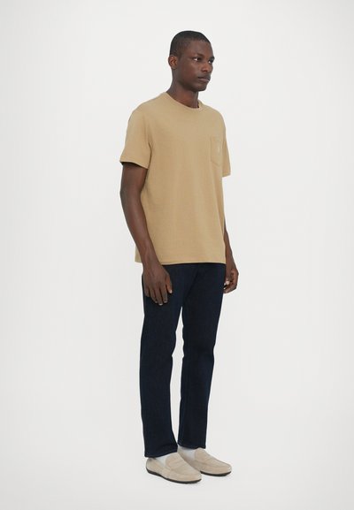 T-shirt beige à manches courtes avec une poche sur la poitrine, associé à un jean bleu foncé et des mocassins beige clair. Design et coupe simples, sans ornements.