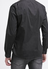 Camicia nera a maniche lunghe con una texture liscia, polsini con bottoni e orlo curvo, mostrata di spalle.