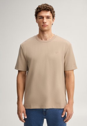 Giovane uomo con capelli ricci che indossa una maglietta beige e jeans blu, in piedi davanti a uno sfondo chiaro e semplice.