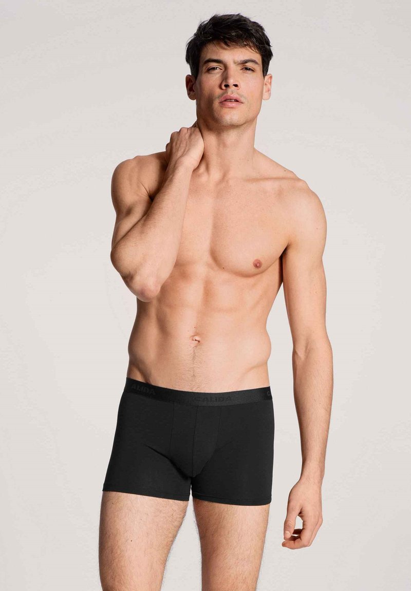 Zwarte boxer shorts met een zachte textuur en een waistband met logo. Het model poseert met een blote bovenlichaam en toont de pasvorm en het ontwerp van het ondergoed.