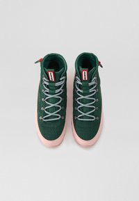 Zapatillas altas verdes con parte superior de tejido texturizado, suela de goma rosa, destacadas con cordones en negro y blanco y una marca en rojo.