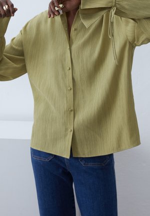 Persoon draagt een licht olijfgroene blouse met textuur, knopen en een strikdetail, gecombineerd met blauwe spijkerbroeken.